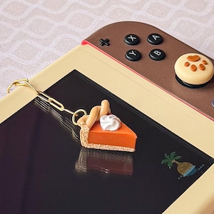 May include: A miniature orange pumpkin pie slice charm with a white whipped cream dollop, attached to a gold-toned paperclip-style chain. The charm rests on a beige and brown handheld gaming console featuring a small palm tree graphic.