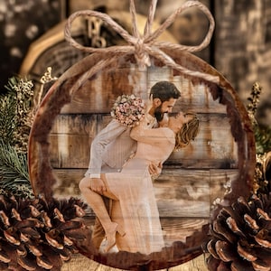 May include: A round wooden ornament featuring a printed photograph of a couple embracing. The ornament has a natural bark edge, a jute twine bow at the top, and is decorated with pine cones. The couple is posed against a rustic wood plank background.