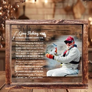 K&ouml;nnte beinhalten: Ein rechteckiges Holzschild mit dem aufgedruckten Bild einer Person mit roter Kappe, die einen Fisch h&auml;lt. Der Text lautet Gone Fishing und enth&auml;lt ein Gedicht &uuml;ber das Angeln. Der Hintergrund zeigt eine rustikale Holzmaserung mit Angelhaken.