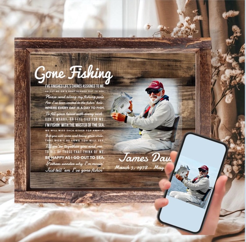 K&ouml;nnte beinhalten: Ein rechteckiges Gedenkschild mit Holzrahmen, rustikalem Holzmaserungshintergrund, dem Text Gone Fishing, einem Gedicht und einem Foto einer Person, die einen Fisch h&auml;lt. Der Name James Dav und Daten stehen unter dem Foto.