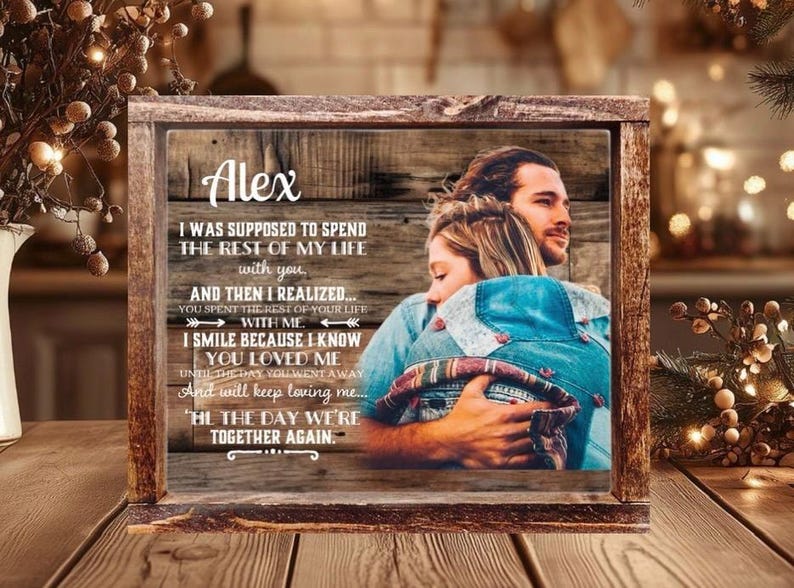 May include: A rectangular wooden frame containing a printed image of a couple embracing. The print features a rustic wood-grain background with the name Alex and a memorial quote about love and loss. The frame has a dark, weathered wood finish.