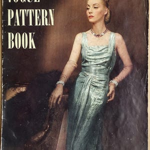 Peut inclure: Une couverture de livre de patrons Vogue vintage représentant une femme en robe bleu clair avec un collier de bijoux. La couverture est brune avec des rayures verticales et le texte "Vogue Pattern Book" en blanc. Le texte "Décembre-Janvier 1937-38" et "Prix 35 cents" est également visible.