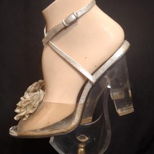 May include: A silver high-heeled sandal with a clear block heel and a strap around the ankle. The sandal features a large, decorative flower on the side and is displayed on a clear stand. The shoe is approximately 4 inches high.