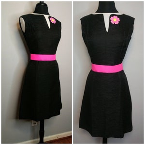 May include: A black sleeveless dress with a pink belt and a pink flower brooch. The dress has a textured fabric and a v-neckline.