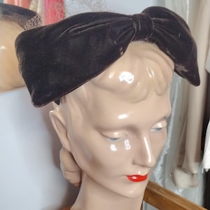 Puede incluir: Un sombrero de lazo de terciopelo marrón oscuro. El sombrero se exhibe en una cabeza de maniquí con lápiz labial rojo. El lazo es grande y se asienta sobre la cabeza. El sombrero es de estilo vintage.