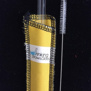 May include: A yellow fabric sleeve with black stitching holds a clear glass drinking straw. A white cleaning brush is next to the sleeve.