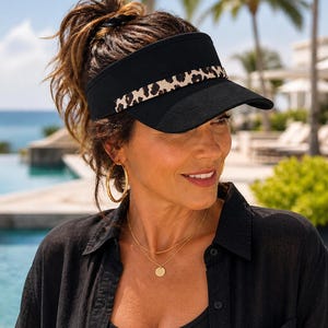 May include: A black visor with a leopard print band. The visor is worn by a person with brown hair and gold hoop earrings. The person is wearing a black shirt and a gold necklace. The background includes a pool and palm trees.
