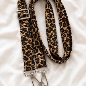 May include: A leopard print phone strap with silver-colored metal hardware. The strap is brown and black with a repeating animal print pattern. The strap has a buckle and two metal clasps for attaching to a phone case.