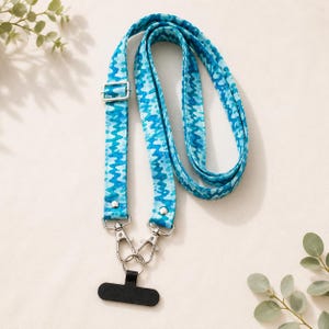 May include: A blue and white patterned lanyard with silver-colored metal hardware. The lanyard has a black phone holder and is designed to be worn around the neck. The pattern is a repeating wave-like design.