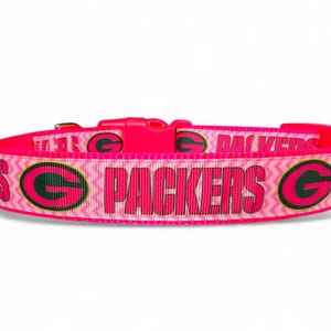 May include: A vibrant pink dog collar with the word "PACKERS" in pink lettering. The collar displays the team's logo and a zig-zag pattern. The buckle is also bright pink. The collar is suitable for a dog.