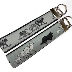 May include: Two keychains with black and white patterned fabric. One keychain features a wolf and sheep, the other features a sheepdog and sheep. Both keychains have silver key rings.