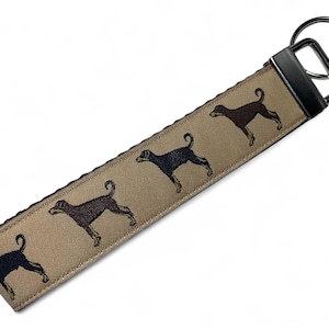 May include: A tan fabric key chain with a black metal key ring. The key chain has a repeating pattern of black and brown silhouette images of Doberman Pinschers.