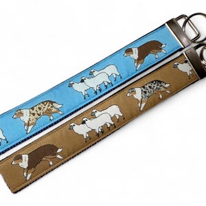 May include: Two keychains with metal rings. One is light blue with a pattern of white sheep and a brown and white dog. The other is tan with white sheep and a speckled dog.