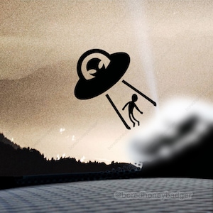 May include: A black silhouette of a UFO with a beam of light shining down on a small alien figure. The image is set against a blurry background of a sunset over a forest.