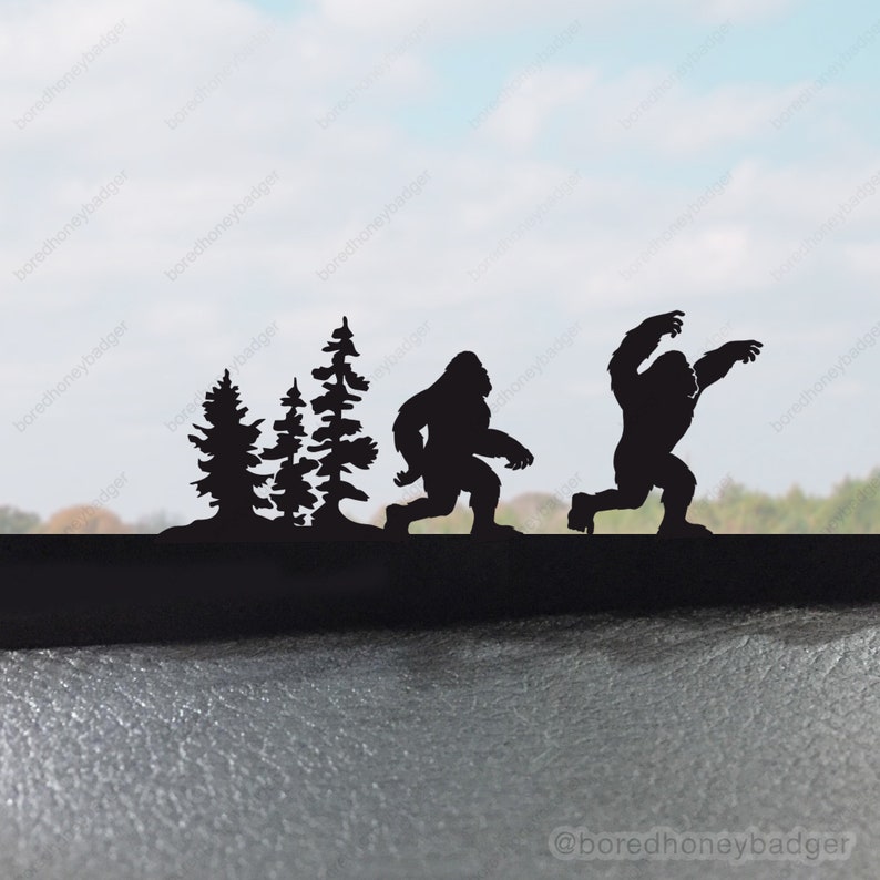 May include: Silhouette of two Bigfoot figures walking through a forest. The figures are black and are set against a blurry background of trees and a blue sky.