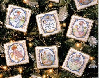 Op de afbeelding: Een verzameling vierkante, kruissteek kerstversieringen. Elk ornament toont een andere bijbelse scène of figuur, omlijst met een blauwe rand en gouden afwerking. De ornamenten hangen aan een kerstboom.