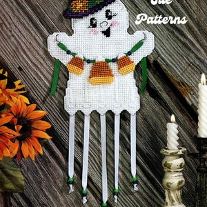 Pode incluir: Um fantasma branco com um chapéu roxo e uma fita verde segurando uma corda de milho doce laranja e amarelo. O fantasma está sorrindo e tem olhos brilhantes. O fundo é uma superfície de madeira com uma vela e girassóis.
