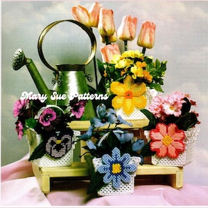 Puede incluir: Una colección de arreglos florales artesanales en cestas blancas tejidas, exhibidos en una pequeña estantería de madera. Los arreglos presentan varias flores coloridas, incluyendo tulipanes, pensamientos y margaritas. También hay una regadera verde. El texto "Mary Sue Patterns" es visible.
