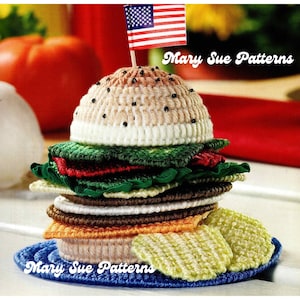 May include: A crocheted hamburger with a small American flag on top, surrounded by a blue plate and yellow and white potato chips. The image includes the text "Mary Sue Patterns".
