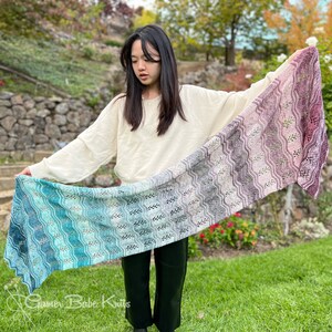 May include: A person wearing a white sweater holds a long, colourful knitted scarf with a wavy pattern. The scarf is a gradient of blue, green, and purple colours. The scarf is made with a delicate lace stitch.