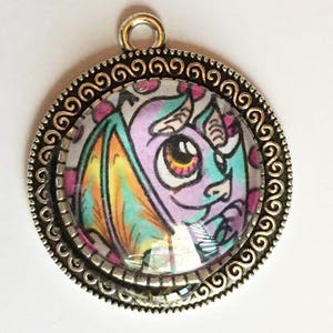 May include: A silver pendant with a round glass cabochon featuring a colorful cartoon dragon with large eyes. The pendant has a decorative silver border with a swirling pattern.