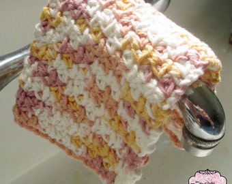 Puede incluir: Una manopla de crochet cuadrada con un patrón de hilo jaspeado en tonos blanco, rosa suave y amarillo pálido. La tela tiene un diseño de puntos repetitivos y texturizados, y está colgada sobre un grifo metálico. Un logo dice The Joy of Crochet Patterns.
