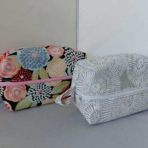 May include: Two rectangular fabric pouches with side handles and zippered closures. One pouch features a black background with a colorful floral pattern in pink, blue, and yellow. The second pouch has a light gray and white circular pattern.