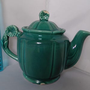 May include: A vintage, emerald green ceramic teapot with a lid and spout. The teapot features a decorative handle and a floral design on the lid. The teapot is approximately 8 inches tall and is suitable for serving tea.