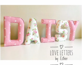 Puede incluir: Letras de tela rosa y blanca que deletrean el nombre "DAISY". Las letras están decoradas con lunares blancos y un patrón floral. Las letras están sobre una superficie blanca. El texto "LOVE LETTERS by Esther" está debajo de las letras.