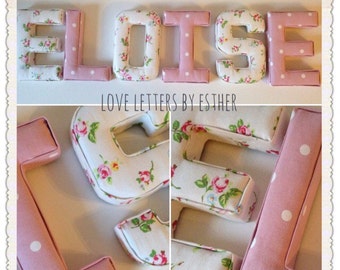 Puede incluir: Letras de tela rosa y blanca que deletrean el nombre "Eloise" con un patrón floral y lunares blancos. Las letras están hechas a mano y tienen un efecto 3D. La imagen también muestra un primer plano de la letra "E" y la letra "L".