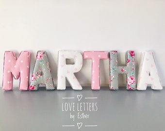 Puede incluir: El nombre "MARTHA" está escrito en letras grandes y coloridas. Cada letra está hecha de tela y tiene un patrón diferente. Las letras son rosas, blancas y grises con diseños florales y de lunares. Las letras están dispuestas en una fila sobre una superficie gris. El texto "LOVE LETTERS by Esther" está impreso debajo de las letras.