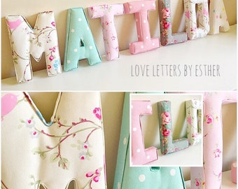 May include: A set of fabric letters spelling out the name "MATILDA" in various floral and polka dot patterns. The letters are made of soft, padded fabric and are perfect for a nursery or child's room. The letters are arranged in a line on a white wall, with the text "LOVE LETTERS BY ESTHER" below them.