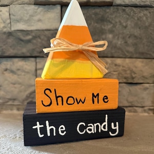 May include: A wooden Halloween decoration featuring a candy corn design. The candy corn is atop two stacked blocks. The top block is painted orange with the words "Show Me" in black. The bottom block is black with "the candy" in white.