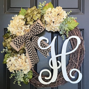 May include: A grapevine wreath adorned with cream and green hydrangeas, greenery, and a black and tan chevron ribbon. A white cursive letter "H" is centered on the wreath, suitable for a front door.