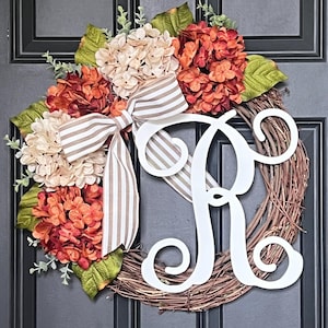 May include: A grapevine wreath featuring a large white script letter R, accented with clusters of cream and burnt orange hydrangea flowers, green leaves, and a beige and white striped fabric bow tied at the top center of the arrangement.