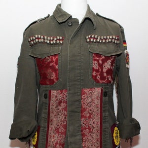 Op de afbeelding: Een groene jas in militaire stijl met rode en gouden stoffen accenten en zilveren studs. De jas heeft een ritssluiting en meerdere zakken.