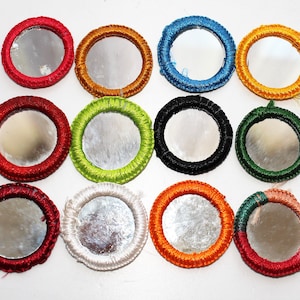 Può includere: Una collezione di dodici piccoli specchi rotondi con cornici ricamate colorate. Le cornici sono in vari colori, tra cui rosso, arancione, giallo, verde, blu, nero e bianco. Gli specchi sono disposti a griglia.