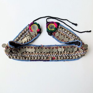 May include: A decorative belt featuring rows of small silver-toned metal coins and blue, white, and black seed beads. The belt includes two circular mirrors framed by purple and green thread, with black braided ties at the top for fastening.