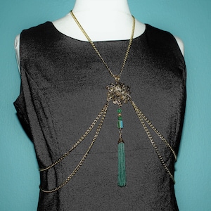 May include: A gold chain body jewelry piece with a green tassel and a floral pendant. The chain is layered and has a delicate design.