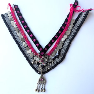 Può includere: Una collana artigianale con base in tessuto nero, finiture argentate e un nastro rosa. La collana presenta un ciondolo argentato con una pietra preziosa rosa e perline d'argento pendenti. Sono attaccate anche piccole monete d'argento.