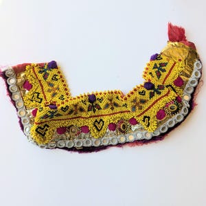 Può includere: Un girocollo tessile curvo caratterizzato da vivaci perline gialle con intricati motivi geometrici, piccoli specchi circolari lungo il bordo esterno, accenti di pon-pon viola e sezioni di tessuto oro metallizzato su una base rosso scuro.