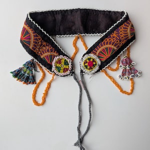 Peut inclure: Un collier textile noir avec des broderies complexes orange, violet et vert. Le collier est orné d'éléments perlés, notamment de petites perles blanches et des ornements circulaires perlés colorés. Des brins perlés orange et des cordons gris pendent du collier.