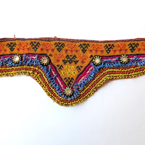 Può includere: Una fascia tessile decorativa con intricate perline nei toni dell'arancione, giallo e blu. Il pezzo è impreziosito da piccoli specchi circolari e motivi geometrici, fissati su una base in tessuto con bordi sfilacciati alle estremità.