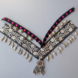 May include: An ornate black and silver embroidered necklace with a V-shaped design. The necklace features silver floral patterns, small silver pendants, and a central pendant with colourful accents. The neckline is trimmed with black fabric and pink ribbon, and silver studs.