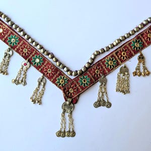May include: A V-shaped decorative belt featuring a deep red fabric band embroidered with green and red floral patterns. The belt is adorned with a row of metallic beads along the top edge and multiple dangling metallic charms and chains.