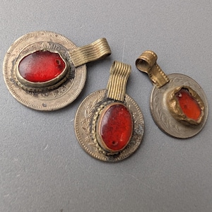 Könnte beinhalten: Drei Vintage-Münzanhänger mit roten Edelsteinzentren. Die Anhänger haben eine silberfarbene Metallbasis mit aufwendigen Details und eine goldfarbene Öse. Die roten Edelsteine sind oval und in einer Lünettenfassung gefasst. Die Anhänger werden auf einer grauen Oberfläche präsentiert.
