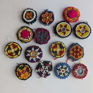 Può includere: Una collezione di piccoli abbellimenti rotondi e perlati in vari colori. I disegni includono motivi floreali e geometrici, con perline nei toni del rosso, giallo, blu e bianco. Alcuni hanno centri a specchio.