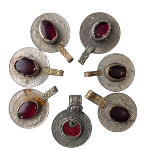 Könnte beinhalten: Eine Sammlung antiker Silberanhänger mit ovalen, tiefroten Edelsteinen. Die Anhänger haben verzierte Designs und kleine Griffe. Einige haben arabische Schriftzeichen. Die Edelsteine sind in Silberfassungen gefasst.