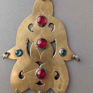 Op de afbeelding: Een goudkleurige metalen hanger in de vorm van een hand, voorzien van drie ronde rode stenen en twee ronde blauwgroene stenen die in het oppervlak zijn gezet. Het metalen oppervlak is versierd met fijne gegraveerde lijnen en kleine lussen.