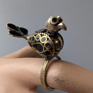May include: A brass bird ring with black and gold detailing. The bird features a detailed design with circular cutouts on its body and wings. The ring band has a textured, rope-like pattern. The bird's head is facing right.